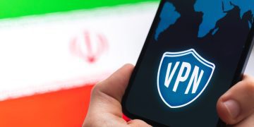 دستی که گوشی هوشمندی با نماد VPN و نقشه جهان را در مقابل پرچم ایران نگه داشته است.