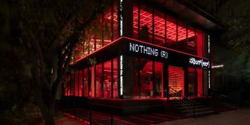 نمای بیرونی فروشگاه رسمی Nothing ® در هند، با معماری مدرن شیشه‌ای، نورپردازی قرمز چشمگیر در فضای داخلی، و لوگوی LED برند Nothing روی نمای ساختمان. طراحی مینیمال فروشگاه با ورودی پلکانی، المان‌های صنعتی و فضای شهری اطراف، هویت بصری متمایز برند را برجسته می‌کند.