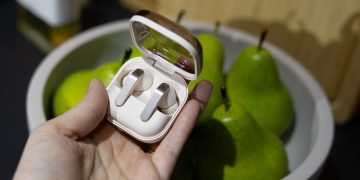 نمای نزدیک از هدفون‌های گلکسی بادز ۴ نقره‌ای داخل کیس شارژ با درپوش شفاف در کنار ظرف گلابی.