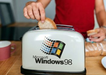 دست فردی در حال قرار دادن نان در توستری که لوگوی بزرگ Microsoft Windows 98 روی بدنه آن نقش بسته است.
