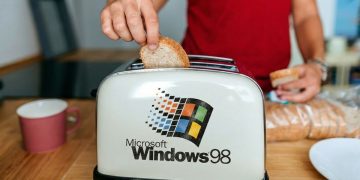 دست فردی در حال قرار دادن نان در توستری که لوگوی بزرگ Microsoft Windows 98 روی بدنه آن نقش بسته است.