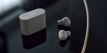 هدفون بی‌سیم Sony XM6 به رنگ نقره‌ای در کنار کیس شارژ روی میز براق.