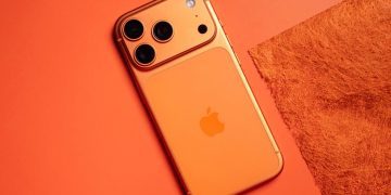 نمای زاویه‌دار از رندر مفهومی یک گوشی آیفون با بدنه نارنجی/برنزی مات، لوگوی اپل در مرکز و ماژول دوربین عقب مستطیلی با سه لنز مجزا، یک فلش دایره‌ای و یک حسگر اضافی؛ گوشی روی پس‌زمینه‌ای هم‌رنگ قرار گرفته است.