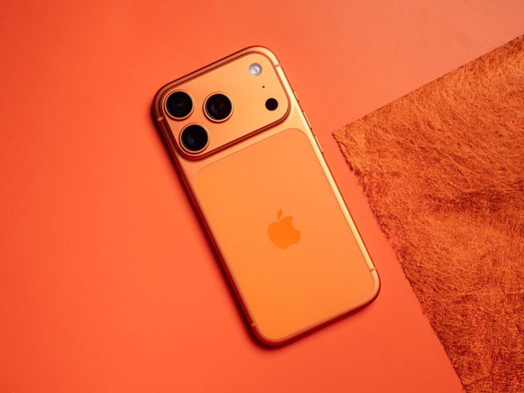 نمای زاویه‌دار از رندر مفهومی یک گوشی آیفون با بدنه نارنجی/برنزی مات، لوگوی اپل در مرکز و ماژول دوربین عقب مستطیلی با سه لنز مجزا، یک فلش دایره‌ای و یک حسگر اضافی؛ گوشی روی پس‌زمینه‌ای هم‌رنگ قرار گرفته است.