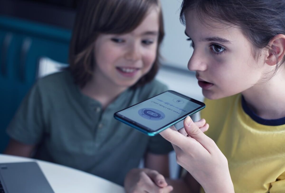 نمای نزدیک از استفاده کودک از تلفن هوشمند در فضای داخلی، در حالی که رابط یک اپلیکیشن یا پیام صوتی روی صفحه گوشی نمایش داده می‌شود؛ تصویری مفهومی درباره تعامل کودکان با فناوری دیجیتال و ابزارهای هوشمند.