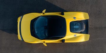 نمای بالا از یک خودروی اسپرت زرد Ferrari با موتور میانی که روی سطح آسفالت قرار دارد و محفظه موتور از پشت شیشه شفاف دیده میشود.