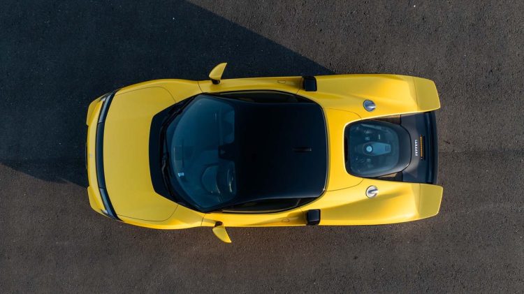 نمای بالا از یک خودروی اسپرت زرد Ferrari با موتور میانی که روی سطح آسفالت قرار دارد و محفظه موتور از پشت شیشه شفاف دیده می‌شود.