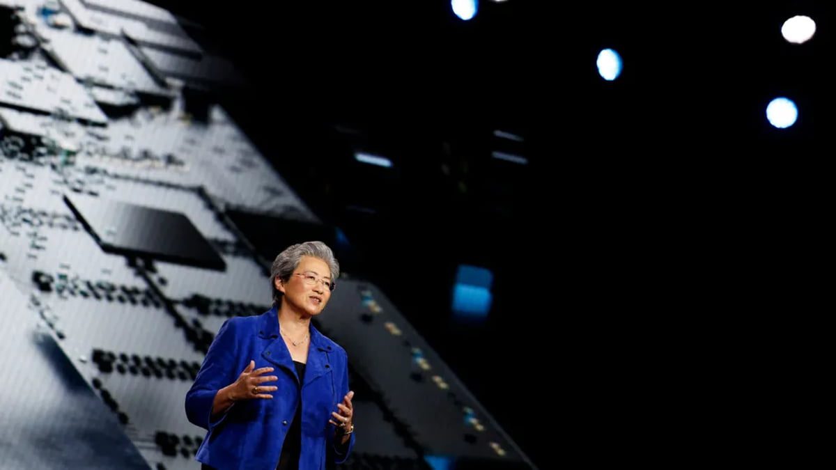 فردی با کت آبی در حال سخنرانی روی صحنه، با حرکات دست توضیح میدهد و پشت سر او تصویر بزرگ یک تراشه و الگوی مدار الکترونیکی نمایش داده شده است