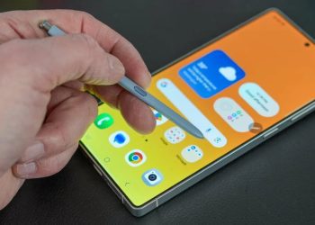 نمای نزدیک از دست کاربر که با قلم S Pen بر روی صفحه‌نمایش یک گوشی هوشمند سامسونگ با پس‌زمینه زرد/نارنجی در حال کار است؛ تأکید بر قابلیت‌های یادداشت‌برداری و دقت قلم روی رابط کاربری One UI