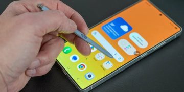 نمای نزدیک از دست کاربر که با قلم S Pen بر روی صفحه‌نمایش یک گوشی هوشمند سامسونگ با پس‌زمینه زرد/نارنجی در حال کار است؛ تأکید بر قابلیت‌های یادداشت‌برداری و دقت قلم روی رابط کاربری One UI