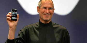 استیو جابز در حالی که نخستین نسل آیفون را در دست گرفته روی صحنه ایستاده؛ صفحهنمایش گوشی با آیکونهای اولیه iOS دیده میشود و پسزمینه شامل لوگوی بزرگ اپل است