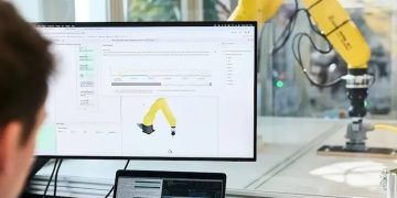 یک بازوی ربات زرد در حال تست در آزمایشگاه و نمایش حرکات آن روی مانیتور کامپیوتر