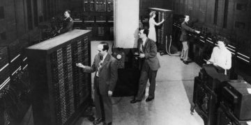 تصویر سیاه‌وسفید تاریخی از یک اتاق کامپیوتر اولیه در دهه‌های میانی قرن بیستم، شامل کابینت‌های بزرگ محاسباتی، پنل‌های پر از سوئیچ و چراغ، و کابل‌های آنالوگ متصل به ماژول‌ها. دو اپراتور در حال کار با پنل‌های کنترلی دیده می‌شوند که مقیاس عظیم و پیچیدگی رایانه‌های نسل اول را نشان می‌دهد.