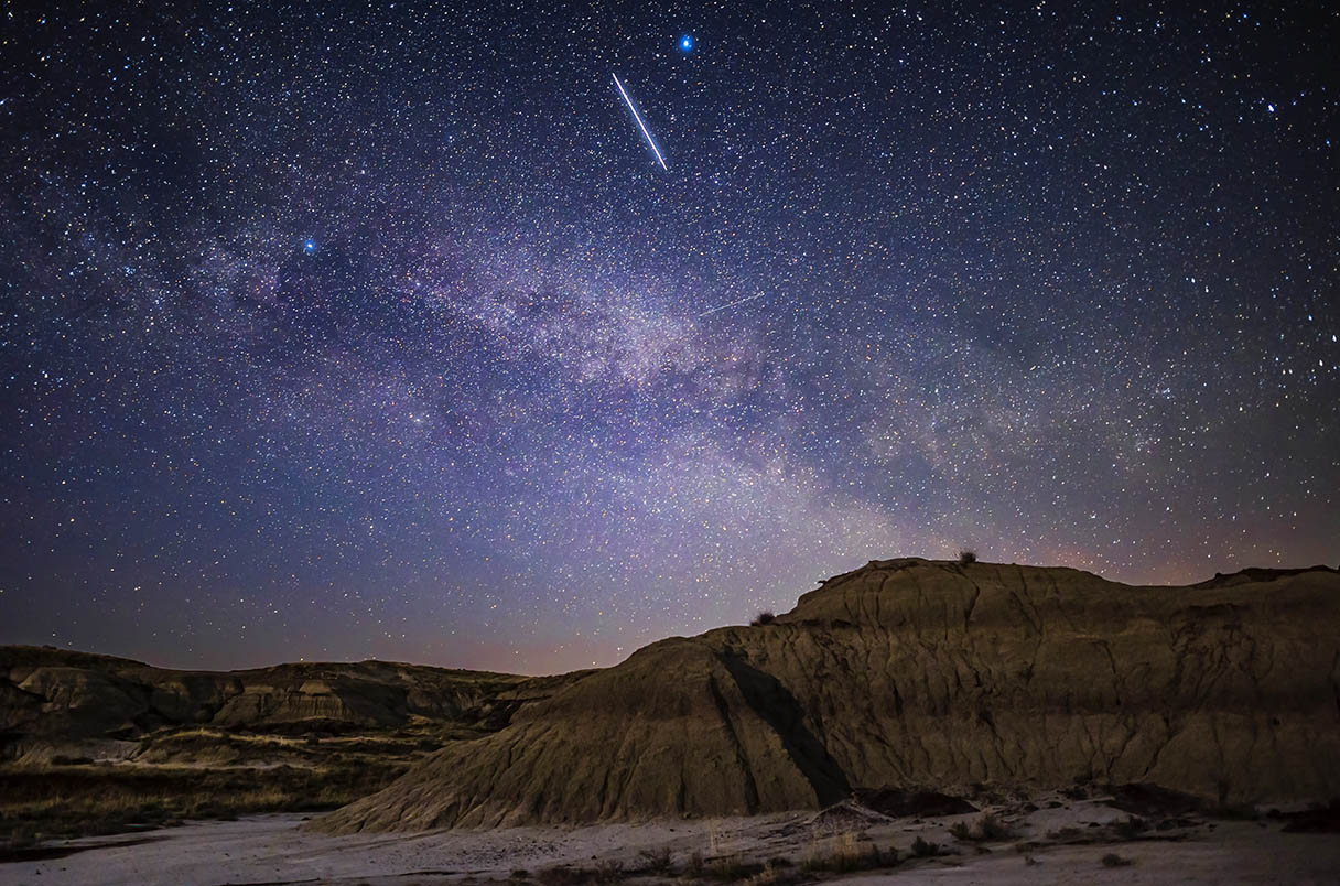 This is a train of closely-spaced Starlink satellites moving to the east over the moonlit landscape of Dinosaur Provincial Park, Alberta. This was on May 15, 2021 at 11:38 p.m. MDT. This group of satellites had been launched from Cape Canaveral earlier that day, at 4:56 p.m. MDT, so less than 7 hours earlier. They were very bright, as shown here, and they appeared as a line or streak in the sky, much as seen here, though the 15-second exposure has turned the array into an even longer streak. It was a remarkable sight, to see a satellite (or satellites in this case) appear not as a star but as a line. To the eye, you could not see them resolved into dots. They are here moving through Lyra below the star Vega. This launch included 52 Starlinks and two other "piggybacked" satellites: the Capella radar satellite, and the NOAA Tyvak-0130 remote sensing satellite. This was the 28th Starlink launch, but is called Starlink 27. This is a single 15-second exposure at ISO 3200 with the Canon R6 and 24mm Sigma Art lens adapted to the R6 and at f/2. Light from the setting waxing crescent Moon provided the illumination. High haze added the star glows.
