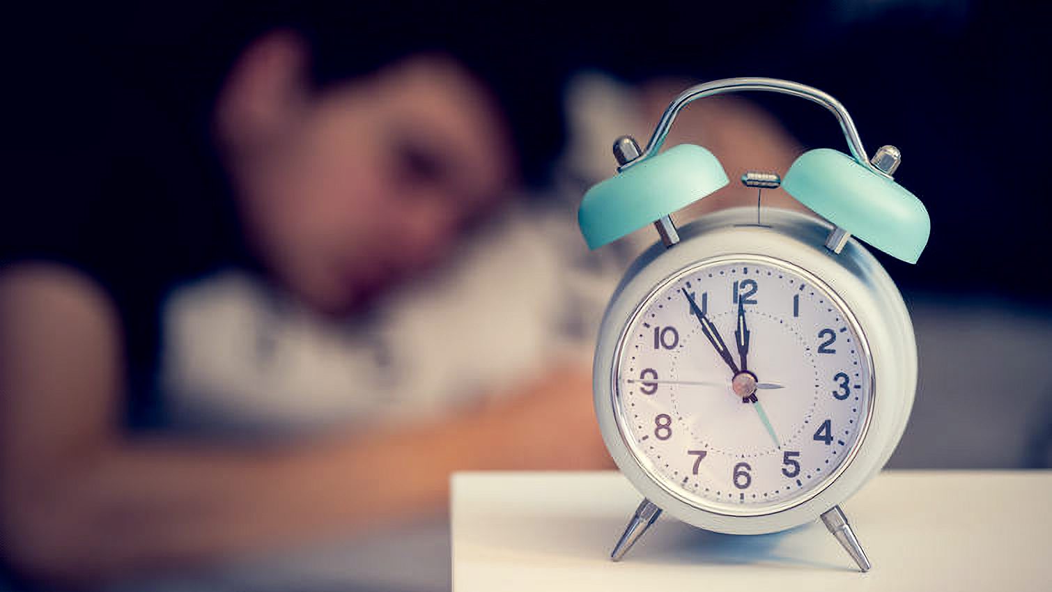 White alarm clock on the bedside table. Young man sleeps in the background.