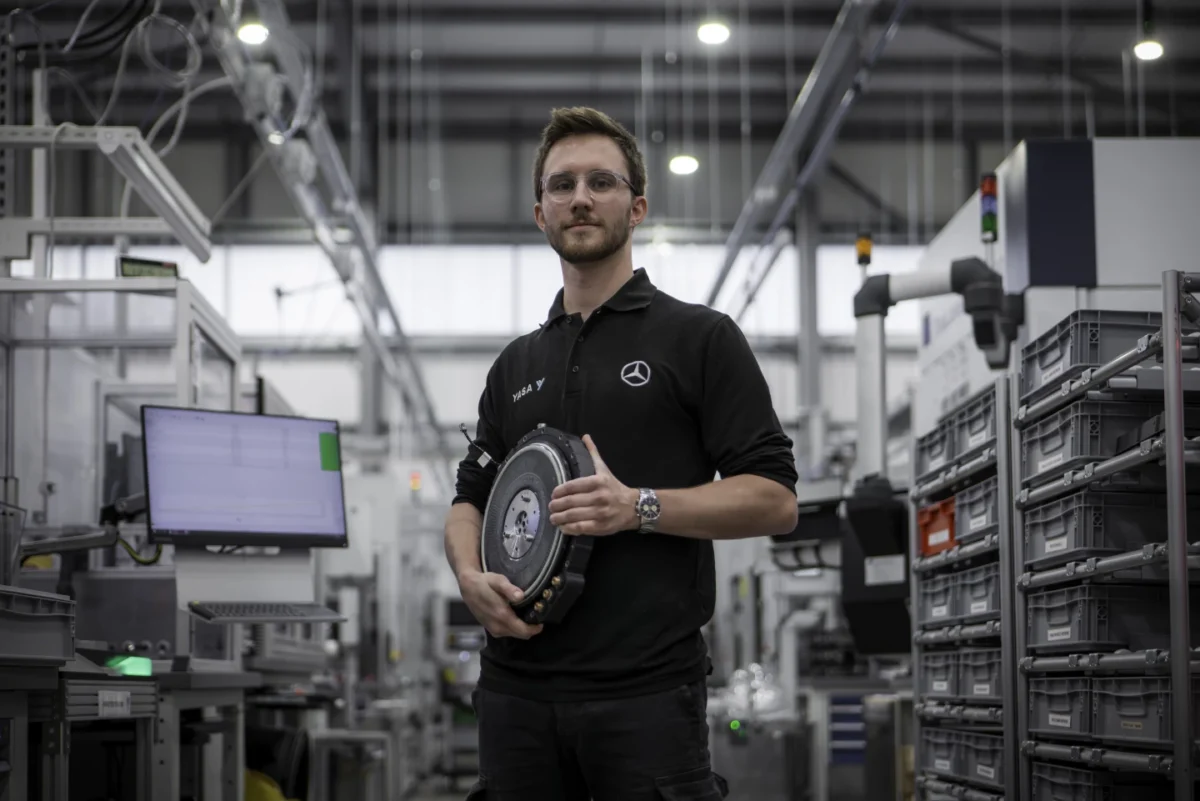 هندس شرکت YASA با لباس مشکی برند Mercedes-Benz در کارخانه در حال نمایش نمونهای از موتور الکتریکی محوری جدید با چگالی توان بالا.