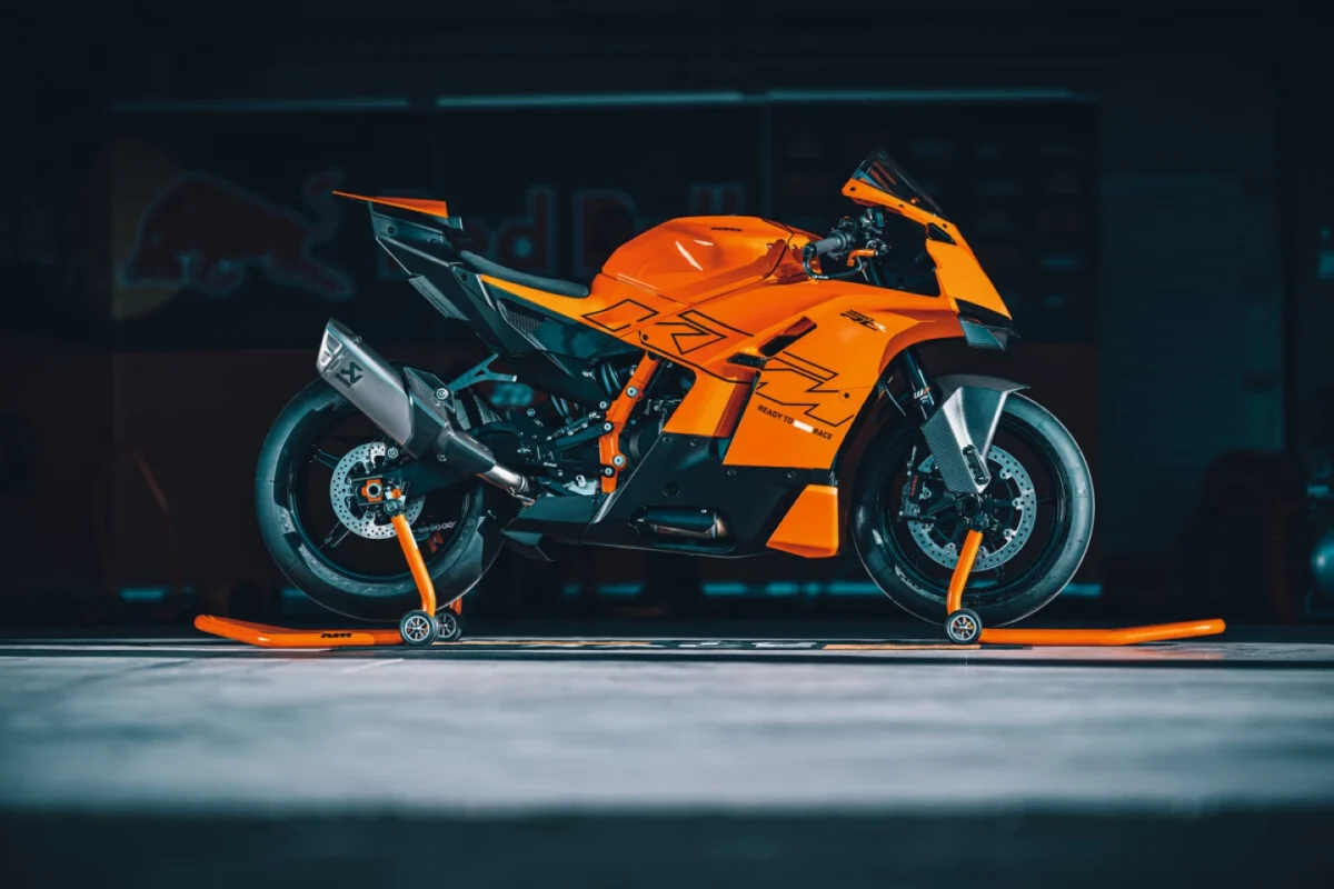 نمای جانبی کامل KTM 990 RC R مدل 2025 روی پایههای نگهدارنده در گاراژ مسابقهای KTM با پسزمینه تاریک.