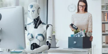 یک ربات انسان‌نما در حال کار با کامپیوتر در یک دفتر در حالی که یک کارمند انسانی در پس‌زمینه وسایل خود را در یک جعبه جمع‌آوری می‌کند.