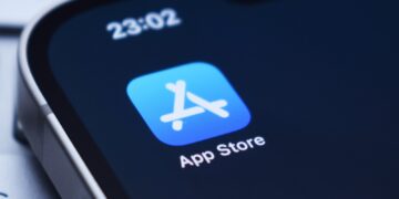 نمای نزدیک از آیکون اپ استور (App Store) بر روی صفحه نمایش یک گوشی هوشمند، با ساعت "23:02" در بالا.