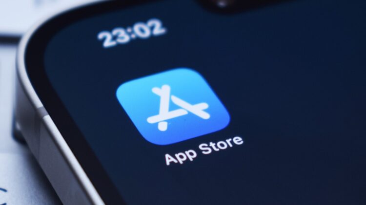 نمای نزدیک از آیکون اپ استور (App Store) بر روی صفحه نمایش یک گوشی هوشمند، با ساعت "23:02" در بالا.