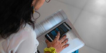 یک زن در حال خواندن یک کتاب درسی است و گوشی هوشمند خود را که نمودار یا داده‌های هوش مصنوعی را روی صفحه نمایش می‌دهد، روی صفحه کتاب قرار داده است.