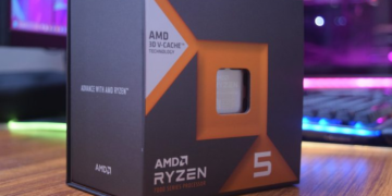 جعبه بسته‌بندی پردازنده AMD Ryzen 5 (7000 Series) با تکنولوژی 3D V-CACHE در یک نمای نزدیک روی یک میز، با پس‌زمینه سیستم کامپیوتری روشن.