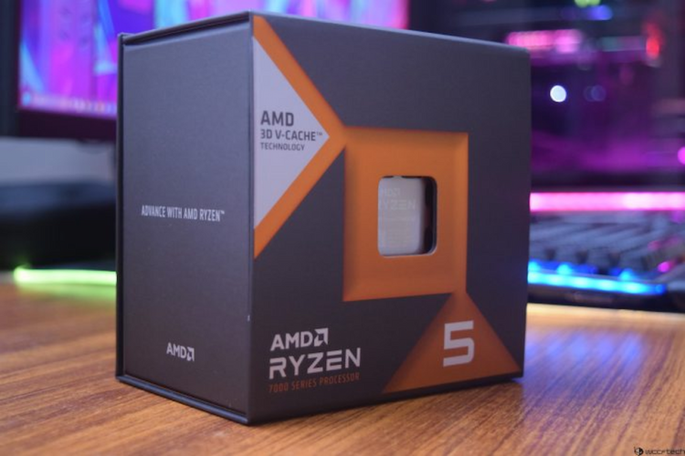 جعبه بستهبندی پردازنده AMD Ryzen 5 (7000 Series) با تکنولوژی 3D V-CACHE در یک نمای نزدیک روی یک میز، با پسزمینه سیستم کامپیوتری روشن.