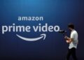 لوگوی Amazon Prime Video به رنگ سفید بر روی یک پس‌زمینه آبی تیره، که یک فرد در حال عبور با تجهیزات دوربین در مقابل آن دیده می‌شود.