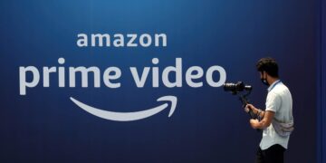 لوگوی Amazon Prime Video به رنگ سفید بر روی یک پس‌زمینه آبی تیره، که یک فرد در حال عبور با تجهیزات دوربین در مقابل آن دیده می‌شود.