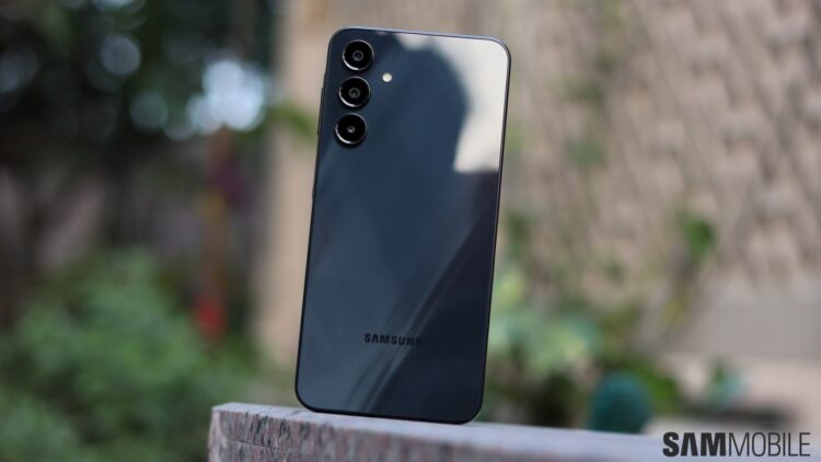 نمای پشت گوشی هوشمند مشکی رنگ سامسونگ (احتمالاً یک مدل Galaxy A یا S جدید) که ماژول دوربین سهگانه با لنزهای جداگانه را نشان میدهد و روی یک سطح سنگی در فضای باز قرار گرفته است.