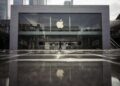 نمای بیرونی یک فروشگاه اپل (Apple Store) با نمای شیشهای بزرگ و لوگوی درخشان اپل در مرکز، که انعکاس آن روی پیادهرو براق دیده میشود.