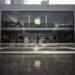 نمای بیرونی یک فروشگاه اپل (Apple Store) با نمای شیشه‌ای بزرگ و لوگوی درخشان اپل در مرکز، که انعکاس آن روی پیاده‌رو براق دیده می‌شود.