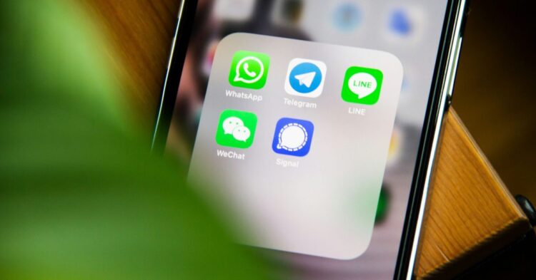 صفحه‌ی اصلی گوشی آیفون با آیکون‌های چندین برنامه پیام‌رسان (WhatsApp، Telegram، LINE، WeChat و Signal) که توسط یک گیاه سبز در پیش‌زمینه قاب شده‌اند.