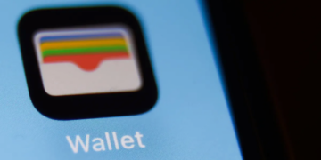 نمای نزدیک از آیکون اپلیکیشن Wallet اپل روی صفحه نمایش یک گوشی با پس‌زمینه آبی روشن.