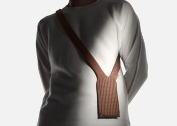 نمای پشتی یک فرد با لباس آستین بلند سفید که "جوراب" یا کیف موبایل iPhone Pocket با بند ضربدری قهوهای (crossbody) را به پشت آویزان کرده است.