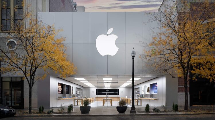 نمای بیرونی یک فروشگاه خردهفروشی اپل (Apple Store) با لوگوی بزرگ اپل روی نمای فلزی و درختان زرد پاییزی در جلوی آن.