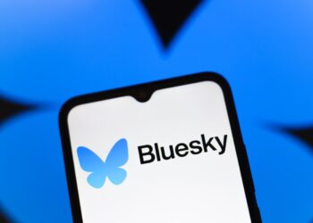 لوگوی Bluesky (پروانه آبی) و نام "Bluesky" بر روی صفحه نمایش یک گوشی هوشمند در مقابل پس‌زمینه آبی تیره.