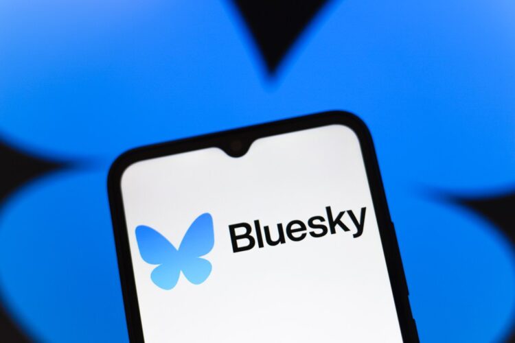 لوگوی Bluesky (پروانه آبی) و نام "Bluesky" بر روی صفحه نمایش یک گوشی هوشمند در مقابل پس‌زمینه آبی تیره.
