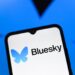 لوگوی Bluesky (پروانه آبی) و نام "Bluesky" بر روی صفحه نمایش یک گوشی هوشمند در مقابل پس‌زمینه آبی تیره.