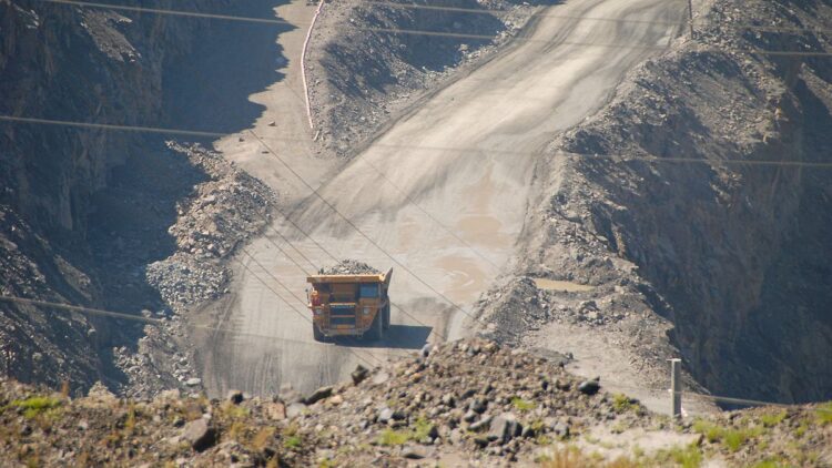 نمای بیرونی معدن روباز بزرگ که کامیون کمپرسی سنگین در حال حمل بار در جاده خاکی پر شیب است.