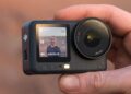 یک دوربین اکشن DJI Osmo Action 6 در دست، که صفحه نمایش جلویی آن یک سلفی از کاربر در محیط بیرون را نمایش میدهد.