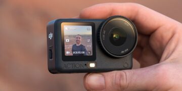 یک دوربین اکشن DJI Osmo Action 6 در دست، که صفحه نمایش جلویی آن یک سلفی از کاربر در محیط بیرون را نمایش میدهد.