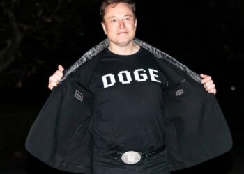 ایلان ماسک در یک محیط تاریک در حال باز کردن کت خود و نشان دادن یک تی‌شرت مشکی که روی آن کلمه پیکسلی "DOGE" نوشته شده است.
