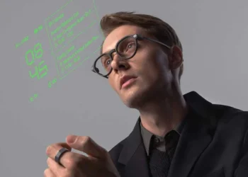 یک مرد جوان در حال استفاده از عینک هوشمند (Smart Glasses) که داده‌های دیجیتالی (ساعت و اطلاعات فهرست) به رنگ نئون سبز را در جلوی چشم او نمایش می‌دهد، و در دستش یک حلقه کنترلر (R1) دارد.