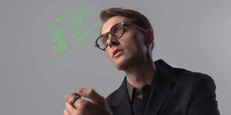 یک مرد جوان در حال استفاده از عینک هوشمند (Smart Glasses) که داده‌های دیجیتالی (ساعت و اطلاعات فهرست) به رنگ نئون سبز را در جلوی چشم او نمایش می‌دهد، و در دستش یک حلقه کنترلر (R1) دارد.