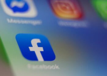 نمای نزدیک از آیکون اپلیکیشن فیس‌بوک (حرف 'f' سفید در مربع آبی) روی صفحه یک گوشی هوشمند با آیکون‌های محو دیگر در پس‌زمینه.