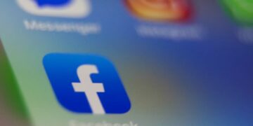 نمای نزدیک از آیکون اپلیکیشن فیس‌بوک (حرف 'f' سفید در مربع آبی) روی صفحه یک گوشی هوشمند با آیکون‌های محو دیگر در پس‌زمینه.