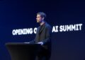 یک مرد در حال سخنرانی در یک کنفرانس (AI Summit) در مقابل یک صفحه نمایش آبی تیره با متن "OPENING OF AI SUMMIT" به صورت برجسته.