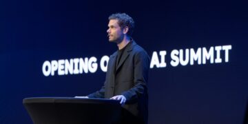 یک مرد در حال سخنرانی در یک کنفرانس (AI Summit) در مقابل یک صفحه نمایش آبی تیره با متن "OPENING OF AI SUMMIT" به صورت برجسته.