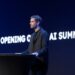 یک مرد در حال سخنرانی در یک کنفرانس (AI Summit) در مقابل یک صفحه نمایش آبی تیره با متن "OPENING OF AI SUMMIT" به صورت برجسته.